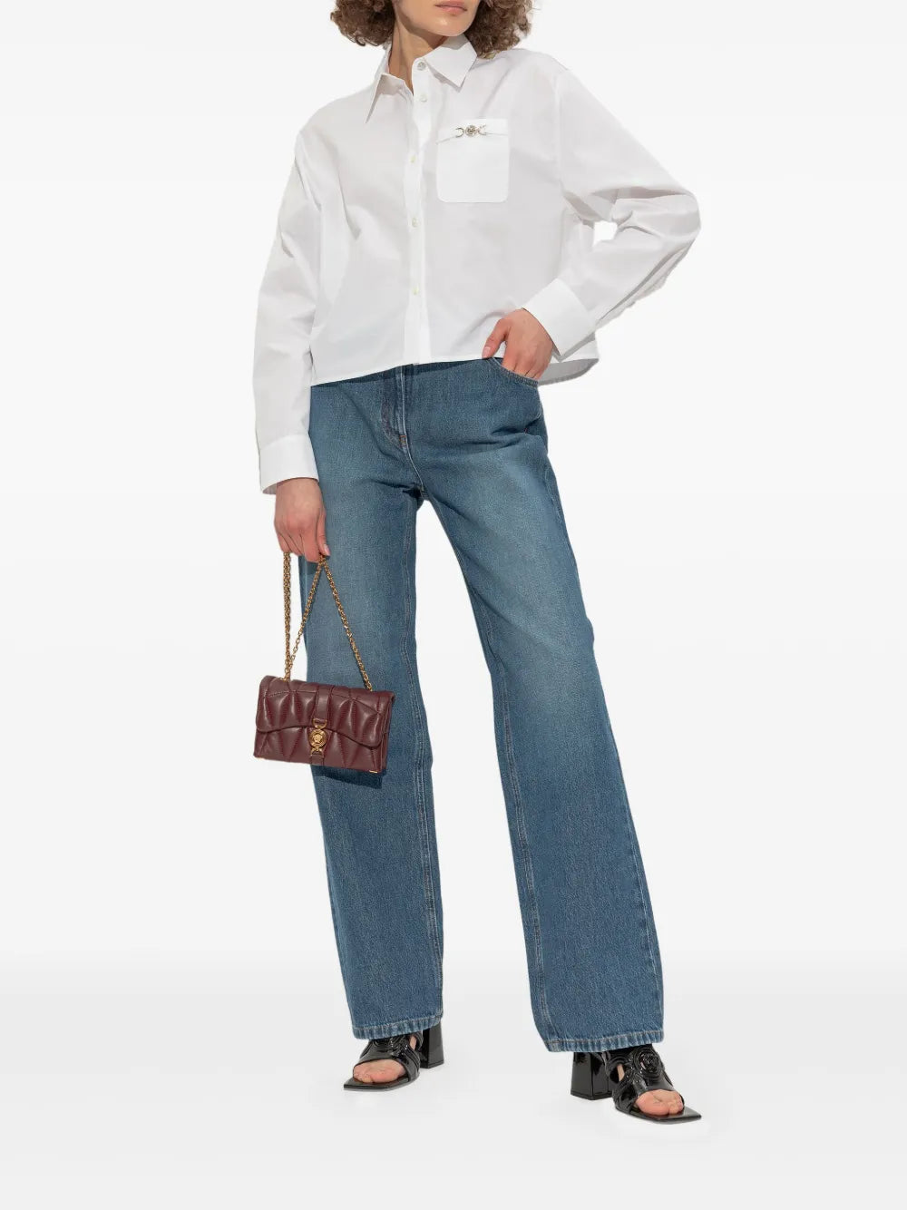 Woman wearing black Versace '95 patent leather mules, white shirt and blue jeans holding a brown Kleio handbag on a white background.